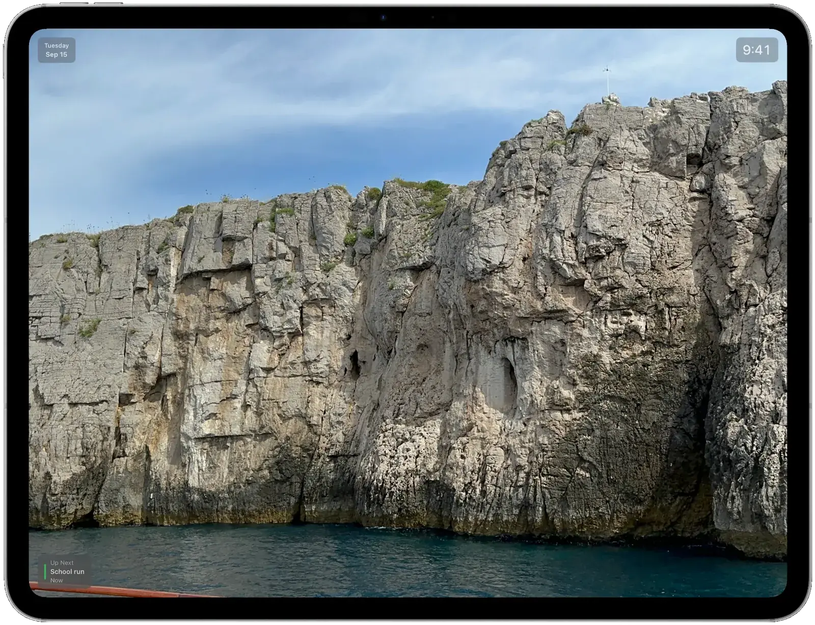 An iPad running Digiframe as a full-screen slideshow of sea cliffs, with the date in the top-left corner, the time in the top-right, and the next calendar event, School run, in the bottom-left.